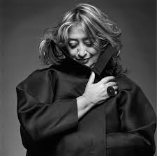 Black-and-white portrait of architect Zaha Hadid, looking down with a soft smile, wrapped in a dramatic dark coat — highlighted by JANGEORGe for her iconic design legacy.