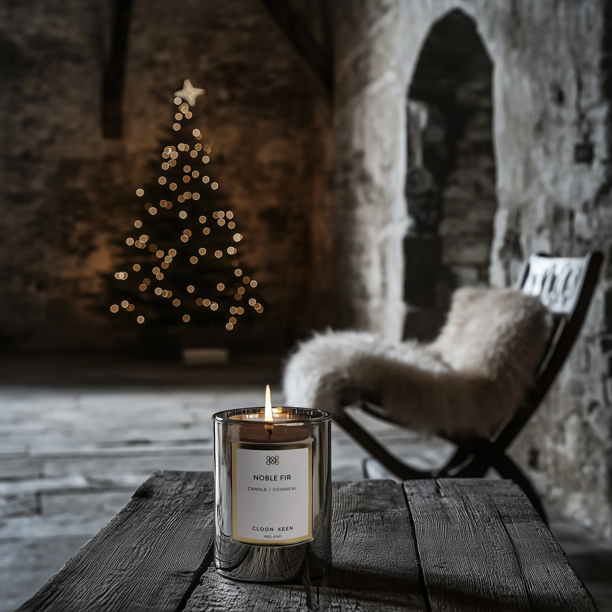 Cloon Keen Noble Fir candle burning on a rustic wooden table in a stone interior with a Christmas tree and fur covered chair in the background, holiday scented candle lifestyle image