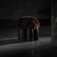 Senufo Ottoman by Arno DeClercq with dark wool seat and Black African Walnut base in a dimly lit space.
