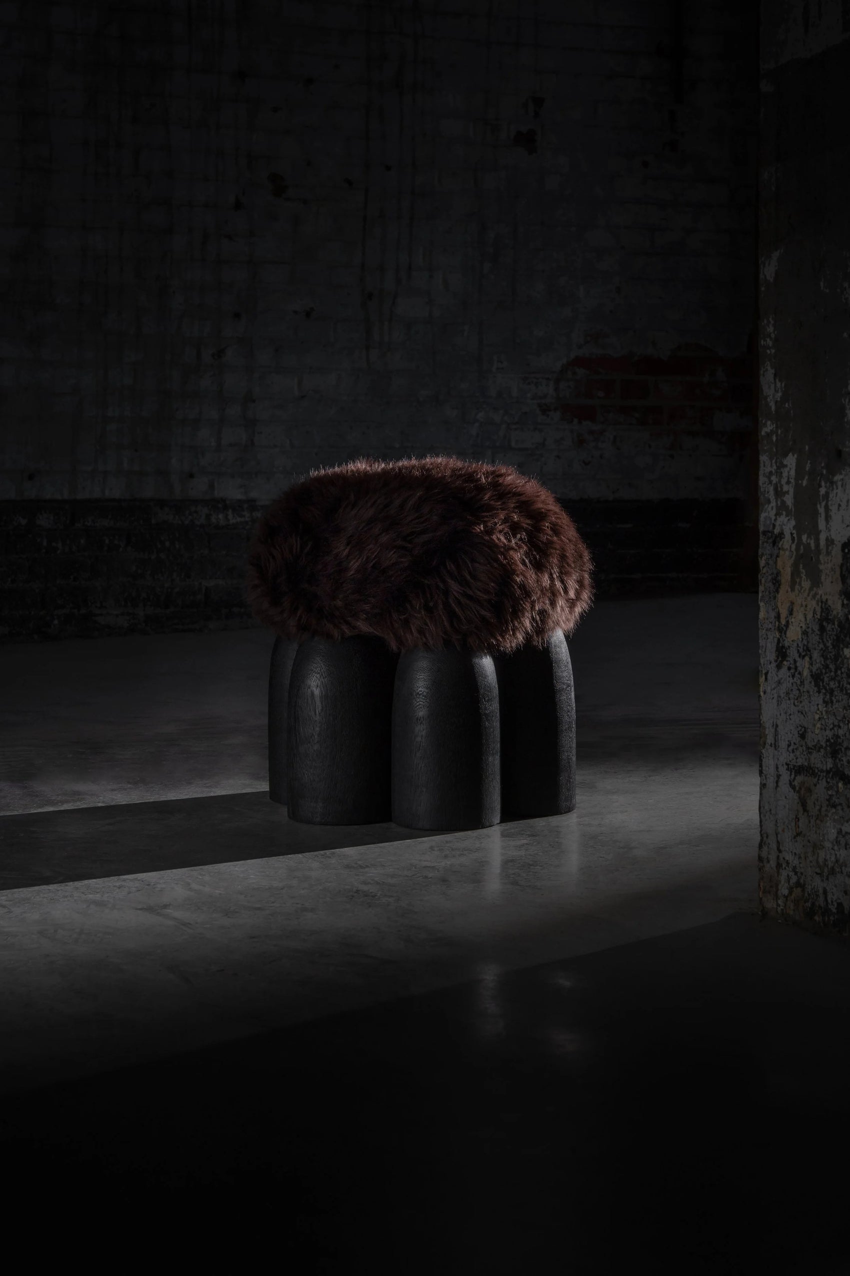 Senufo Ottoman by Arno DeClercq with dark wool seat and Black African Walnut base in a dimly lit space.