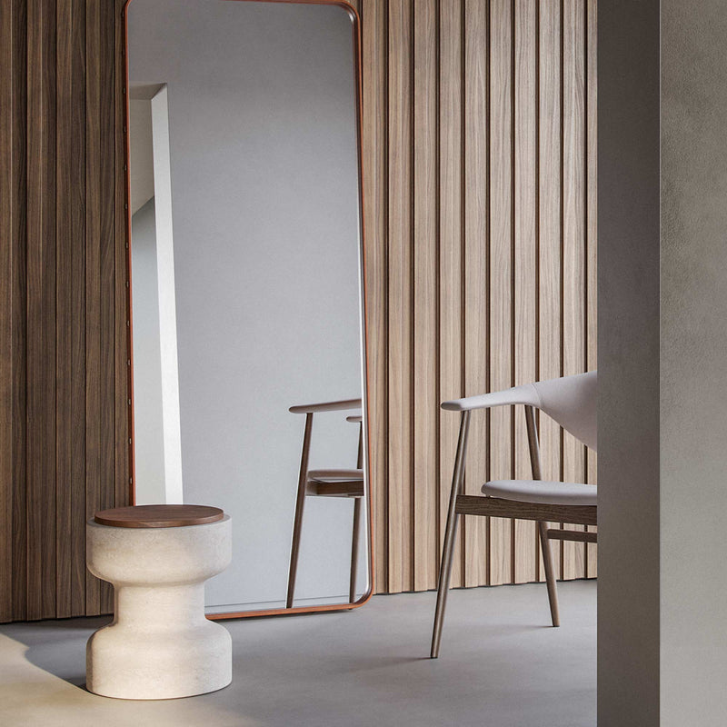 Tivoli stool in Travertino Navona base and Walnut wood top (bottom left). In a room with a mirror and a chair with wood paneled wall in background.