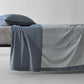 Society Limonta Nim bed sheet in blue denim cotton draping over a bed, highlighting the reversible two-tone color and soft texture.