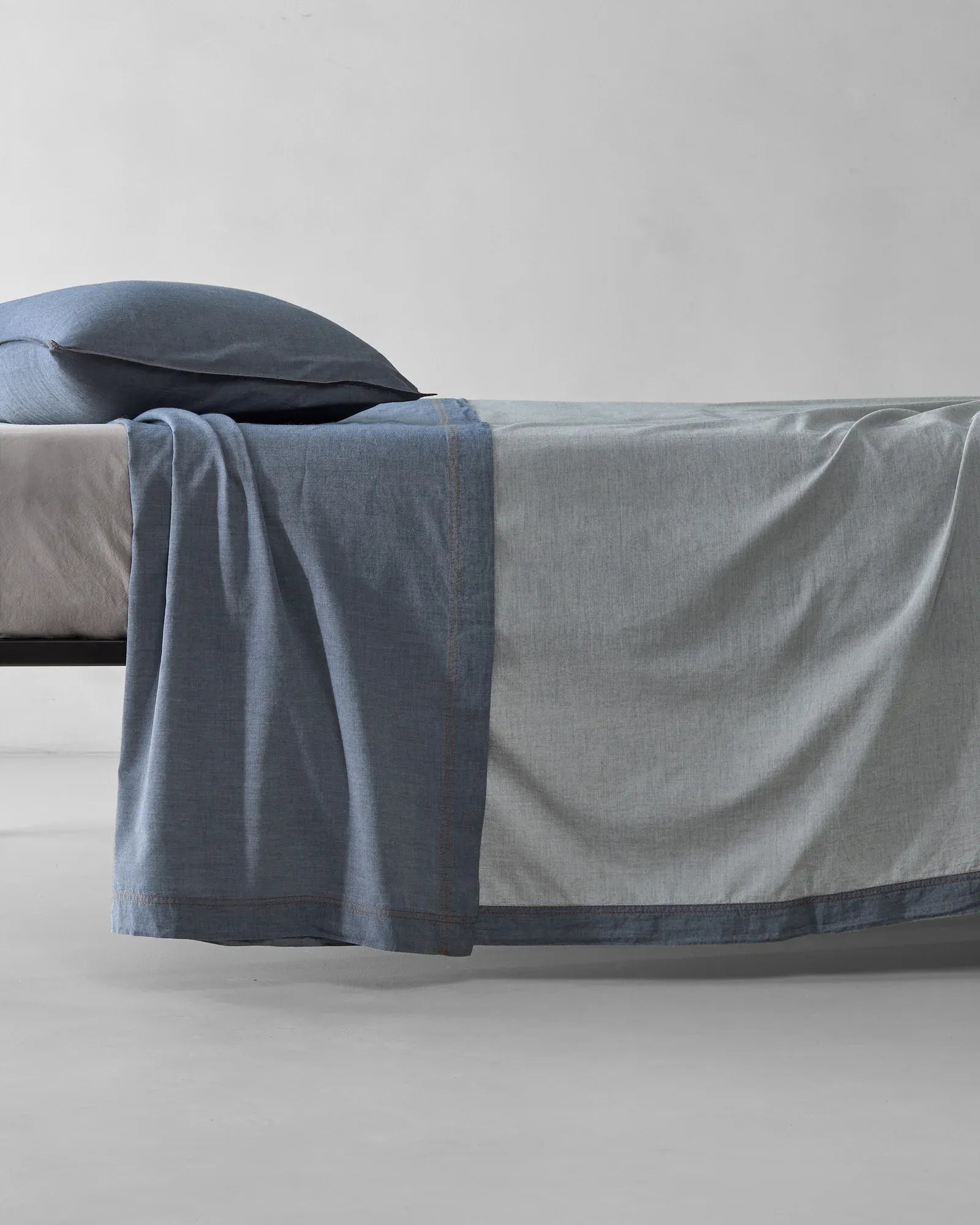 Society Limonta Nim bed sheet in blue denim cotton draping over a bed, highlighting the reversible two-tone color and soft texture.